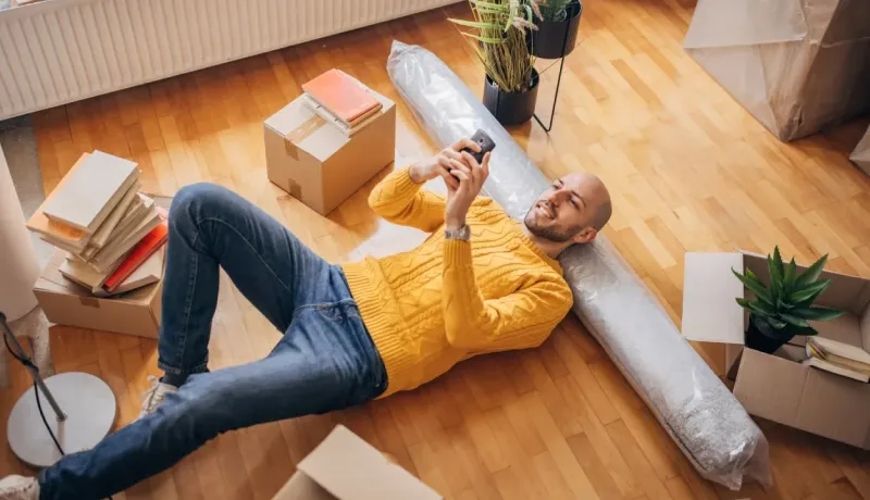 Homme se reposant sur le sol de sa nouvelle maison, entouré de cartons et d'affaires, regardant son téléphone.