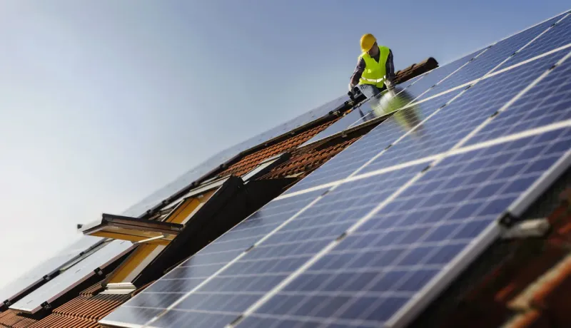 Ouvrier sur un toit, vêtu d'un gilet jaune, fixant des panneaux solaires par un ciel bleu dégagé.