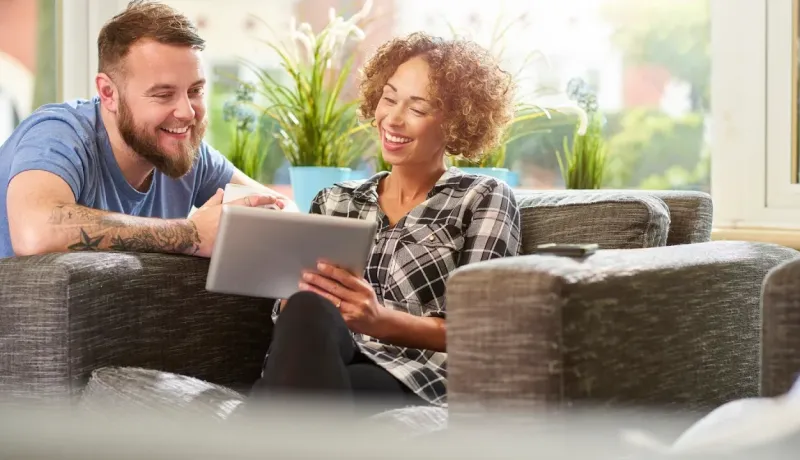 Couple joyeux assis sur un canapé, la femme tenant une tablette numérique, partageant un moment de détente.