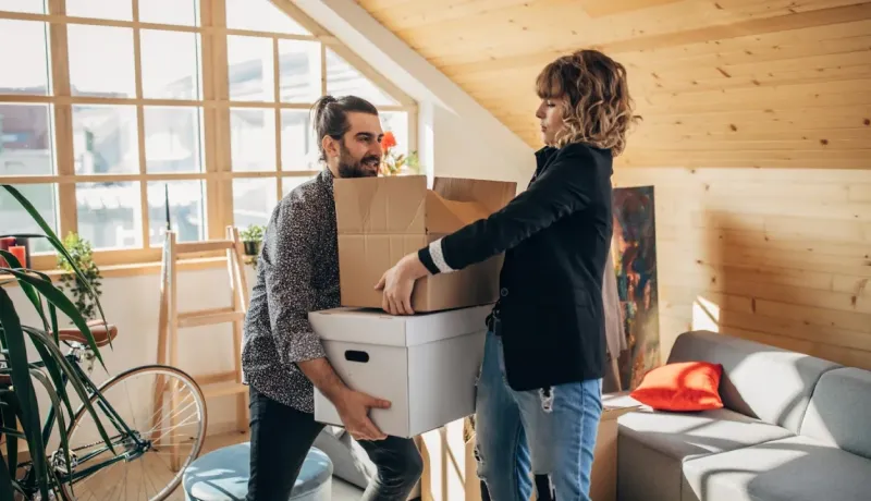 Couple transportant des cartons dans une pièce lumineuse avec un mur en bois et une grande fenêtre, déménagement ou rangement.