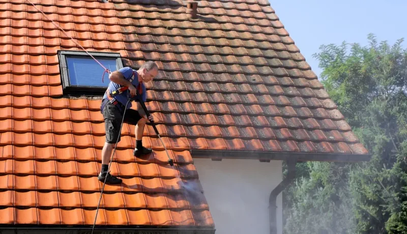 Homme sur un toit de tuiles, sécurisé par un harnais, nettoyant la toiture avec un nettoyeur haute pression.