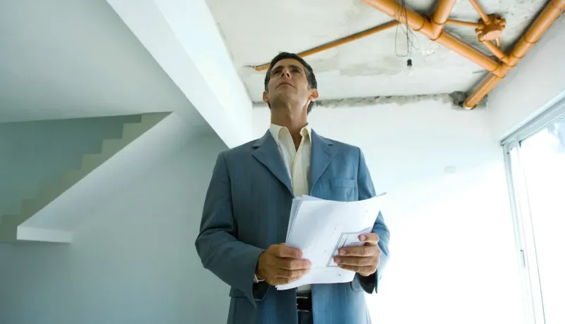 Homme en costume regardant un plafond avec des tuyaux exposés et tenant des plans. Inspection de travaux.