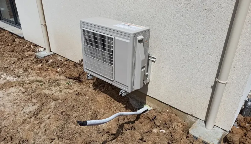 Unité extérieure de climatisation fixée au mur d'une maison sur un terrain non aménagé.