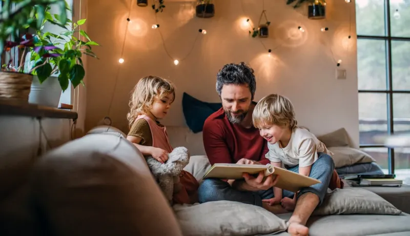 Père souriant lisant un livre à ses deux jeunes enfants sur un canapé, éclairés par des guirlandes lumineuses.