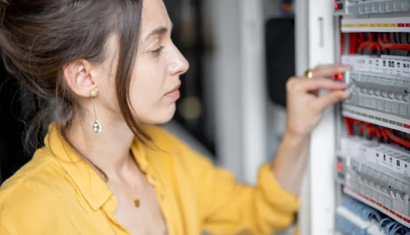 Jeune femme en chemise jaune vérifiant ou réarmant un disjoncteur dans un tableau électrique.