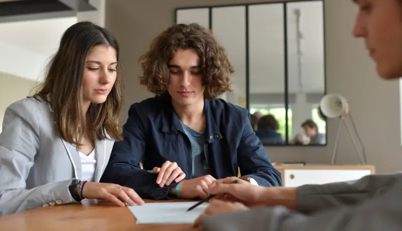 Jeune couple attentif assis à une table en bois, consultant un document avec un conseiller ou agent.