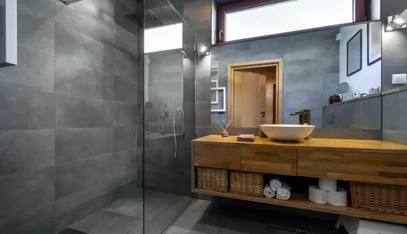 Salle de bain moderne avec douche à l'italienne, carrelage gris, et meuble-vasque en bois avec miroir.