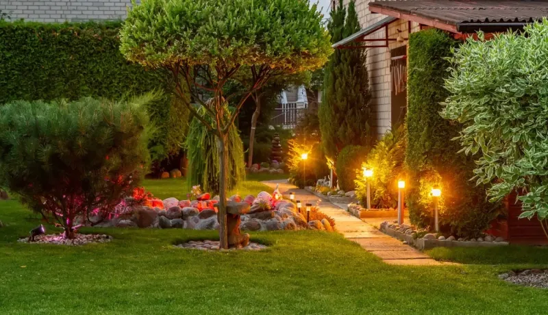 Chemin de jardin éclairé par des lampes d'extérieur le long d'une maison et d'un parterre de verdure.
