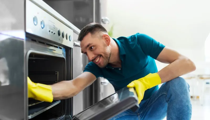 Un homme souriant portant des gants de protection jaunes nettoie l'intérieur d'un four de cuisine.
