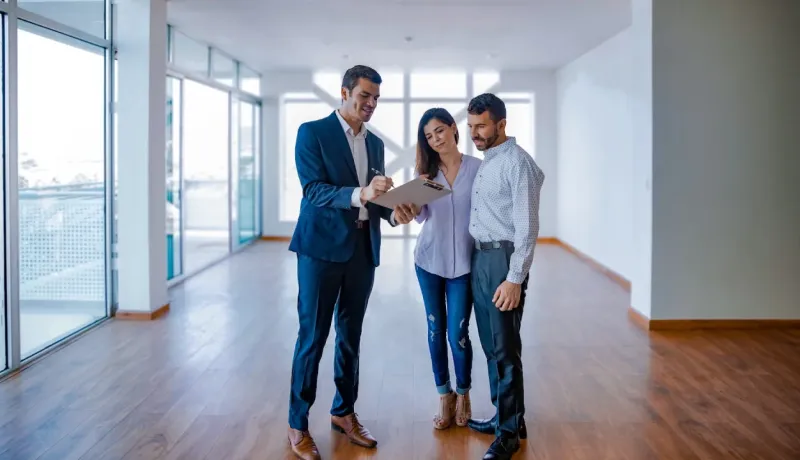 Un agent immobilier en costume discute de documents avec un jeune couple debout dans une grande pièce vide avec des fenêtres.
