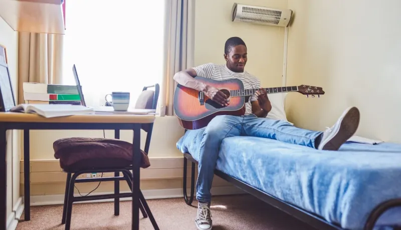 Un jeune homme noir est assis sur un lit dans une chambre d'étudiant, jouant de la guitare acoustique près d'un bureau.