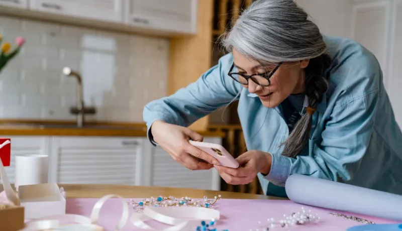 Une femme asiatique aux cheveux gris prend une photo de ses bijoux artisanaux avec un téléphone dans une cuisine.