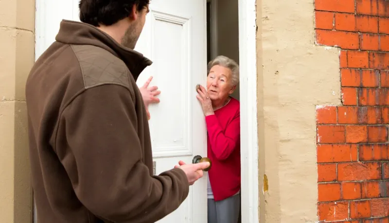 Un homme discute avec une femme âgée qui regarde prudemment par l'entrebâillement de sa porte d'entrée.
