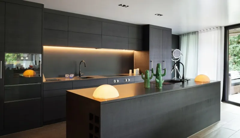 Une cuisine moderne aux armoires foncées et élégantes, avec un îlot central, un éclairage sous armoire et une grande fenêtre.