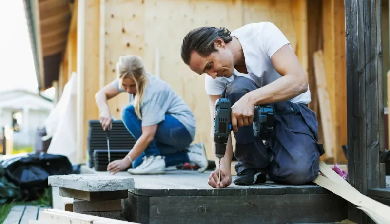 Un couple travaille ensemble pour construire une terrasse extérieure, utilisant des outils comme une visseuse et un tournevis.
