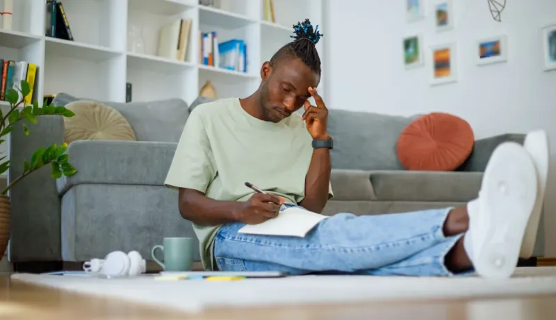 Un jeune homme concentré est assis par terre dans son salon, écrivant dans un cahier.
