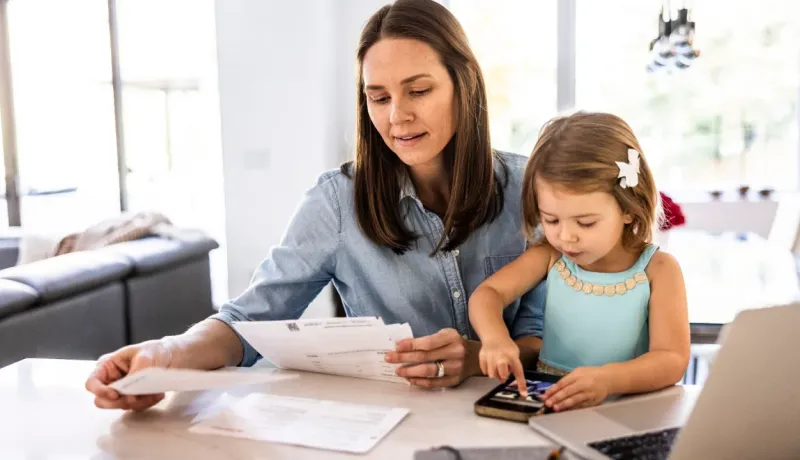 Une mère et sa petite fille sont assises à une table, la mère examinant des documents et l'enfant jouant avec un téléphone.