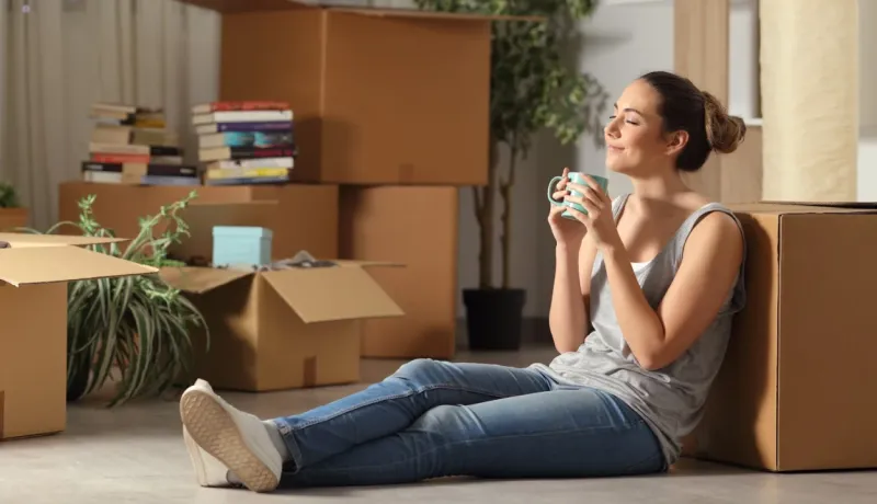 Une jeune femme se détend sur le sol, les boîtes de déménagement empilées autour d'elle, profitant d'une tasse de boisson.