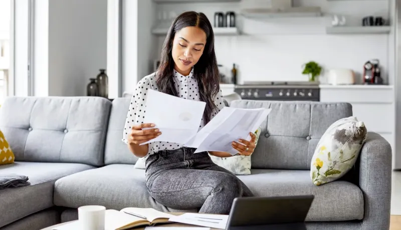 Femme aux cheveux longs assise sur son canapé, consultant des documents financiers et des graphiques.