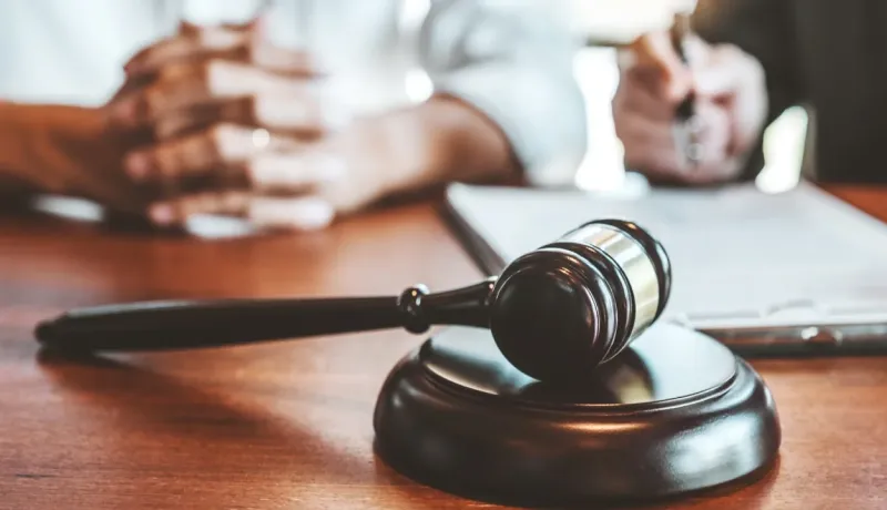 Un gros plan sur un marteau de juge (maillet) posé sur un socle en bois, en face de personnes signant un document juridique.