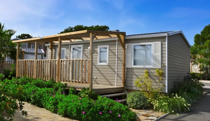 Un mobil-home de loisirs avec terrasse en bois et clôture, entouré de végétation sous un ciel bleu dégagé.