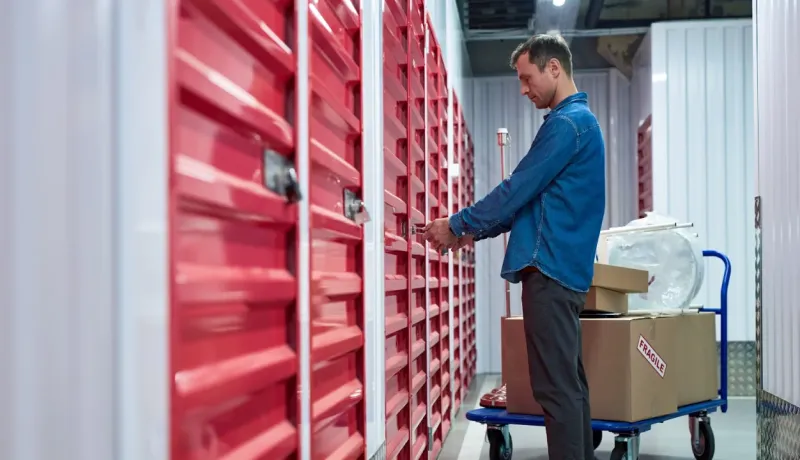 Un homme ouvre le cadenas d'une unité de stockage rouge, avec un chariot de boîtes et d'articles à côté.