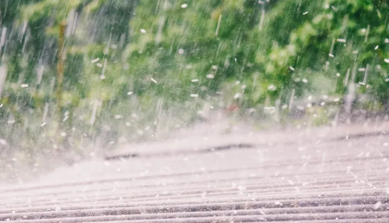Gros plan d'une très forte pluie tombant en gouttes épaisses sur un toit, avec un flou de verdure en arrière-plan.