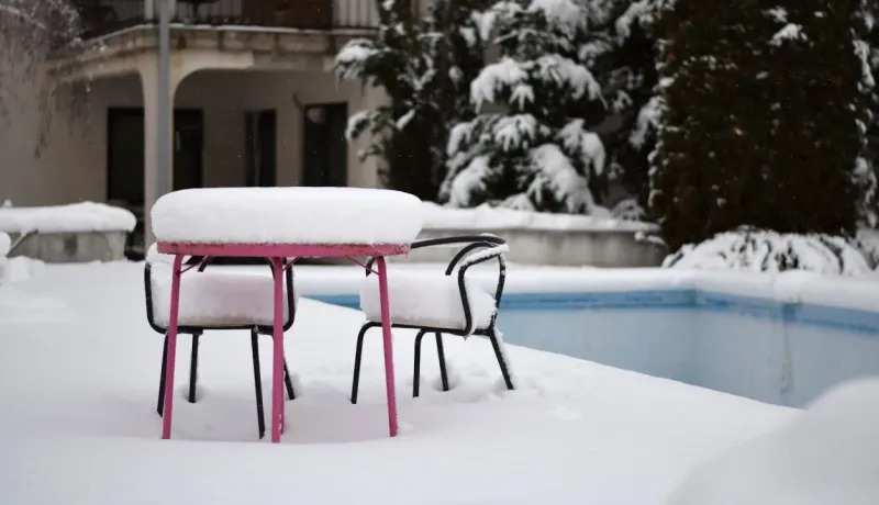 Table de patio rose vif et deux chaises noires totalement enfouies sous une épaisse couche de neige fraîche, près d'une piscine.