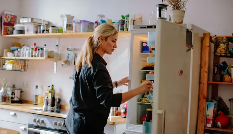 Une femme blonde ouvre et regarde à l'intérieur d'un réfrigérateur ancien dans une cuisine encombrée et bien remplie.