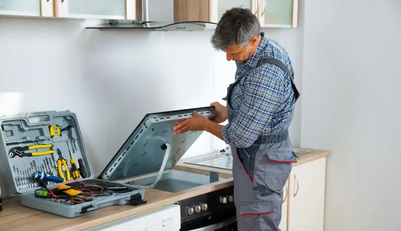 Un réparateur en salopette grise et chemise à carreaux soulève et examine une plaque de cuisson électrique.
