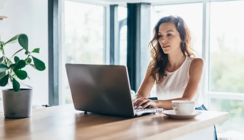 Une femme travaille sur son ordinateur portable, un café à côté, assise à une table en bois près d'une grande fenêtre lumineuse.