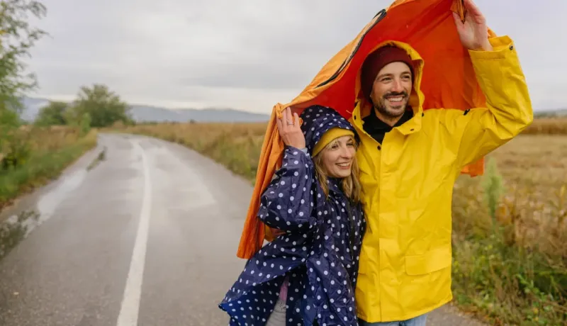 Un couple souriant se protège de la pluie sous un manteau orange sur une route rurale mouillée, vêtus d'imperméables.