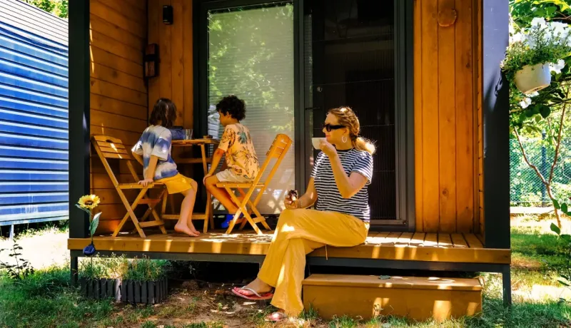 Une femme boit son café à l'extérieur d'une petite maison en bois pendant que deux enfants déjeunent à côté d'elle.
