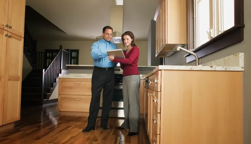 Un agent immobilier et une femme examinent un document dans une cuisine moderne aux armoires en bois clair.