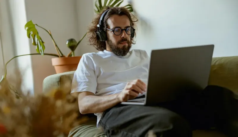 Un homme barbu portant des lunettes et un casque travaille sur un ordinateur portable, assis sur un canapé vert.