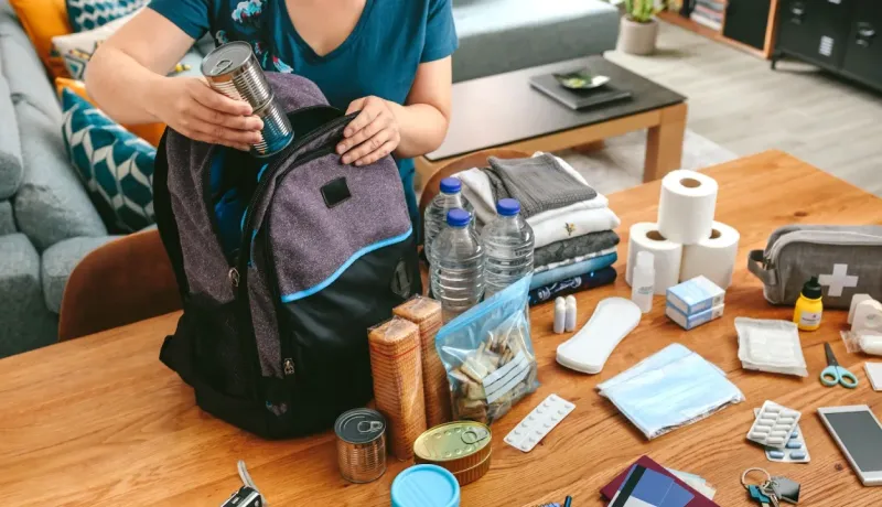 Préparation d'un sac d'urgence avec de la nourriture en conserve, des bouteilles d'eau et des fournitures médicales sur une table.
