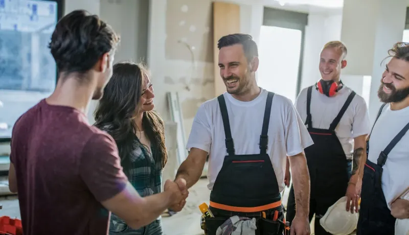 Un contremaître souriant serre la main du client dans une maison en rénovation, le reste de l'équipe est à l'arrière-plan.