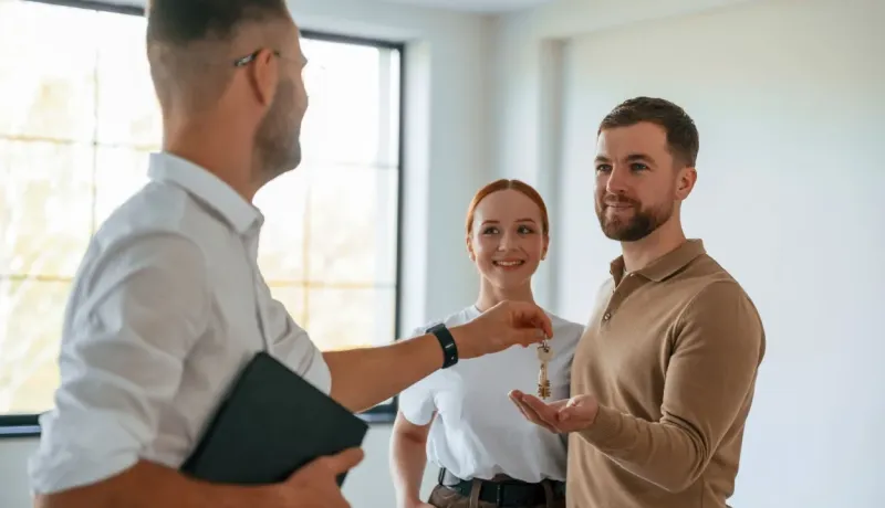Un agent immobilier en chemise blanche tend les clés de maison à un jeune couple souriant et heureux.