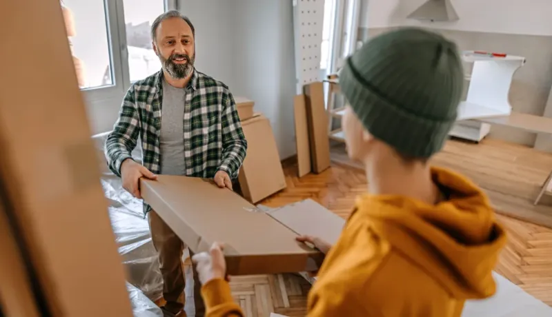 Un père et son enfant assemblent des meubles préfabriqués. L'homme tient un carton dans une pièce en rénovation.