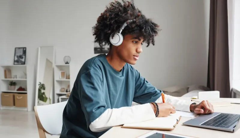 Jeune homme aux cheveux bouclés portant un casque, concentré sur la prise de notes tout en travaillant sur un ordinateur.