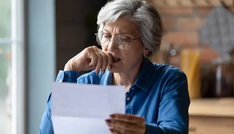 Portrait d'une femme âgée avec des lunettes lisant une lettre ou un document important, visiblement inquiète et pensante.
