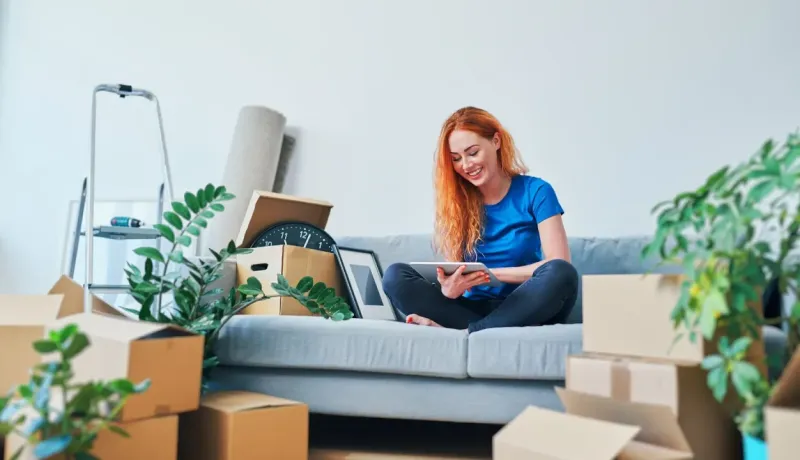 Une femme rousse sourit en utilisant une tablette, assise sur un canapé au milieu de cartons de déménagement et de plantes.