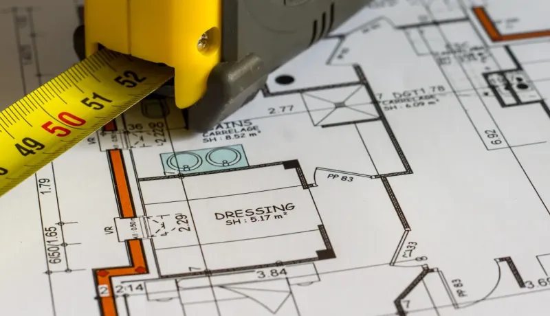 Un mètre ruban jaune et gris est posé sur un plan d'architecte, indiquant des mesures et des détails de construction.