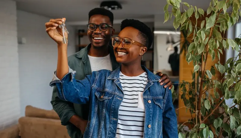 Un jeune couple souriant d'origine africaine, avec des lunettes, tient fièrement en l'air les clés de leur nouvelle maison.