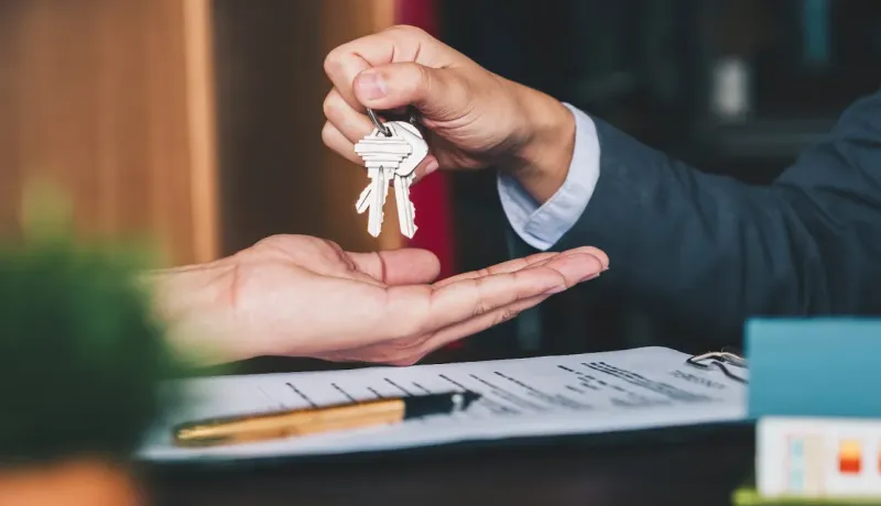 Gros plan d'une main d'agent immobilier remettant des clés à un client, avec un contrat et un stylo-plume sur le bureau.