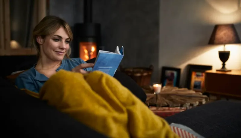 Une femme est détendue sous une couverture jaune, lisant un livre dans un salon sombre et confortable avec un poêle à bois allumé.