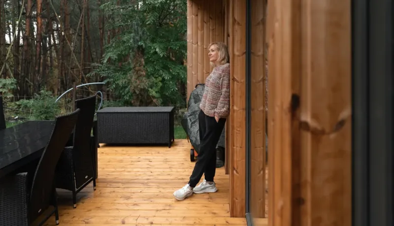 Une femme se détend contre le mur d'une cabane en bois, les yeux levés. Terrasse en bois en premier plan, forêt en arrière-plan.