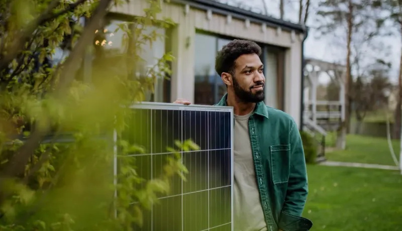Un homme barbu tient un panneau solaire, posant devant sa maison. Symbole de l'énergie verte pour l'habitat.