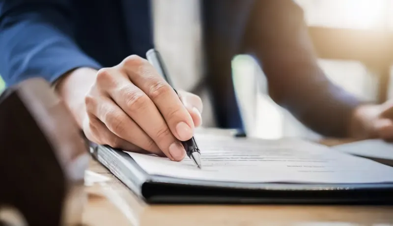 Gros plan sur une main signant un document avec un stylo noir, dans un bureau bien éclairé.