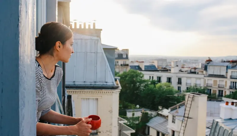 Femme buvant une boisson chaude, regardant le paysage urbain de Paris depuis un balcon ensoleillé.